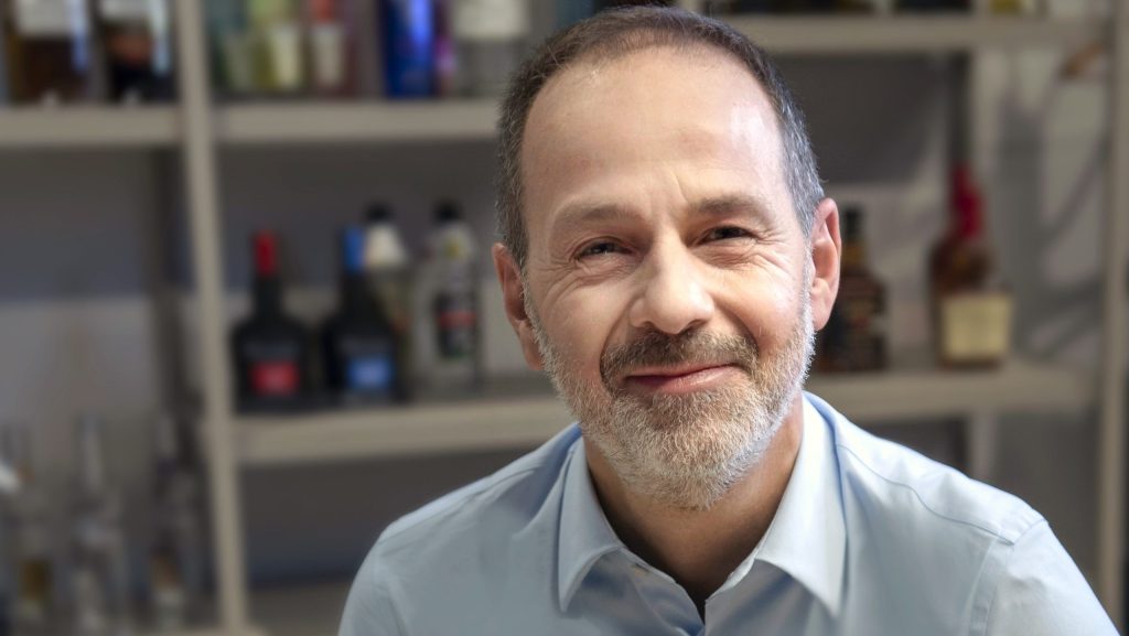 man smiles at camera wearing blue shirt, background of blurred spirits bottles on shelves.