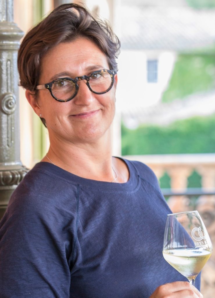 Woman looking at camera, short hair, dark glasses with glass of white wine.